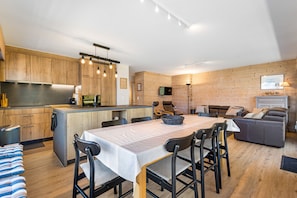 A modern kitchen with wooden cabinets, a dining area with a table and chairs, and a living area with a sofa and a fireplace.