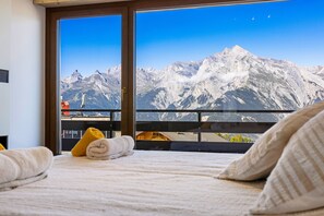 Ein Hotelzimmer mit Bett, Kissen und Blick auf schneebedeckte Berge durch ein großes Fenster.