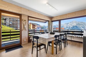 A modern dining area with a large table and chairs, wooden walls, and a view of mountains and a ski lift.