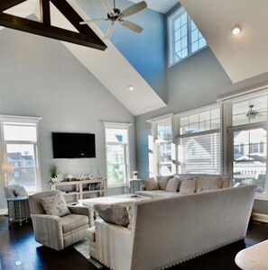 A spacious living room with a high ceiling, large windows, a ceiling fan, and a flat-screen TV mounted on the wall.