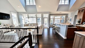 Amplia sala de estar con techo alto, ventanas grandes, cocina con alacenas de madera oscura y una escalera con barandilla blanca.