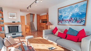 A modern living room with a fireplace, a sofa with red and blue cushions, a wooden coffee table, and a large painting of a snowy mountain scene. - Muverans II F5 - 8 pers - duplex (Haute-Nendaz)