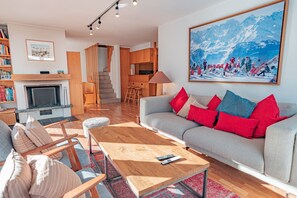 A modern living room with a fireplace, a sofa with red and blue cushions, a wooden coffee table, and a large painting of a snowy mountain scene.