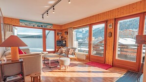 A wooden-floored living room with a sofa, chairs, a TV, and a view of a mountain landscape through large windows. - Muverans II F5 - 8 pers - duplex (Haute-Nendaz)