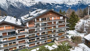 A multi-story hotel with balconies, a ski slope, and snow-covered mountains in the background. - Muverans II F5 - 8 pers - duplex (Haute-Nendaz)