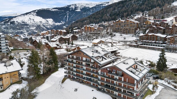 A snow-covered mountain town with multi-story buildings, including a prominent hotel with balconies and a ski resort in the background. - Muverans II F5 - 8 pers - duplex (Haute-Nendaz)