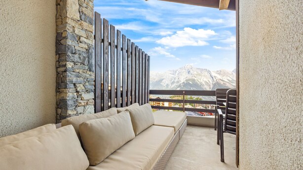 A balcony with a stone wall, a beige sofa, and a view of mountains. - Les Hauts-de-Nendaz A C5 - 5 pers - ski In&Out (Haute-Nendaz)