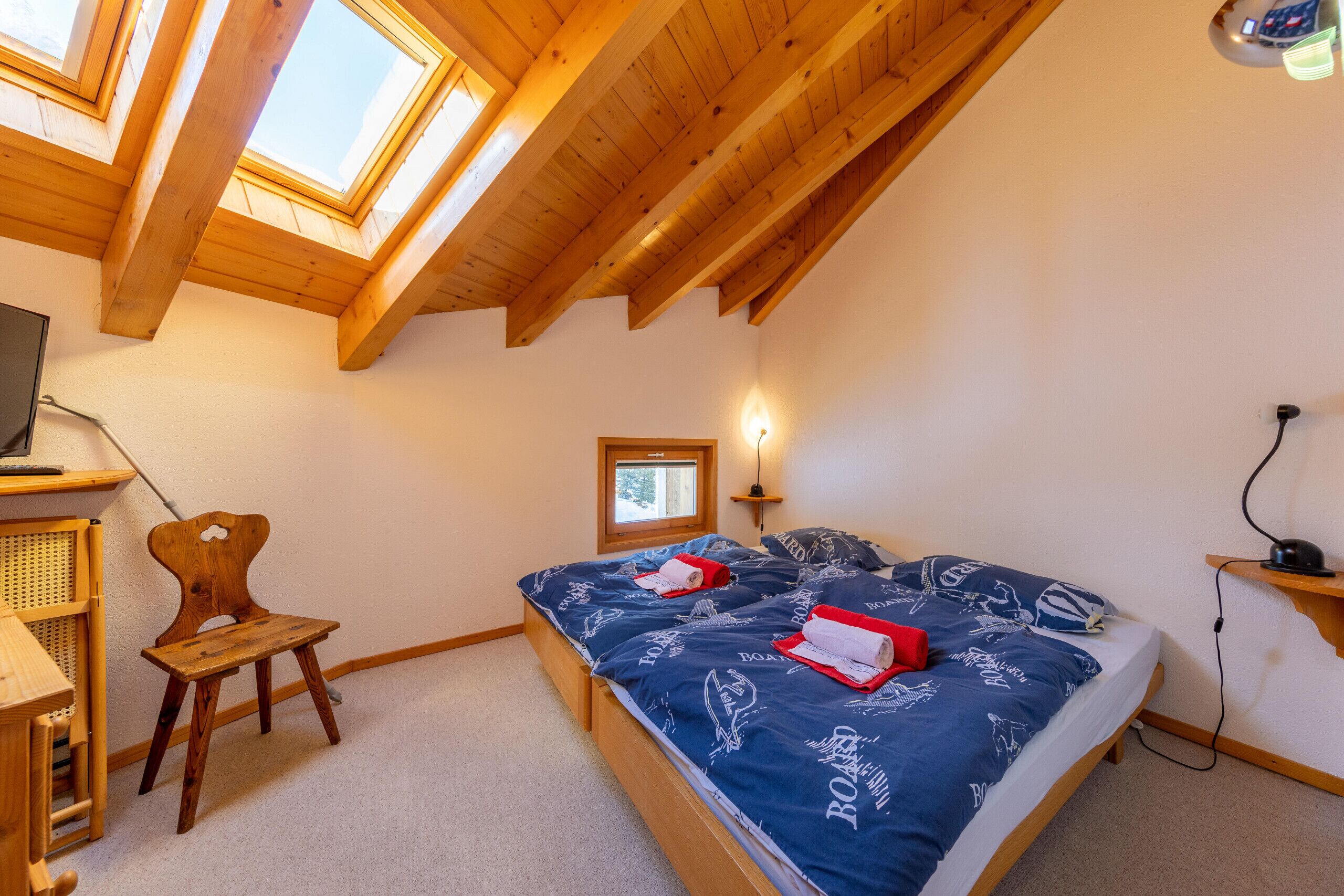 A bedroom with a sloped ceiling, skylights, a bed with a blue bedspread, a wooden chair, and a desk with a computer.