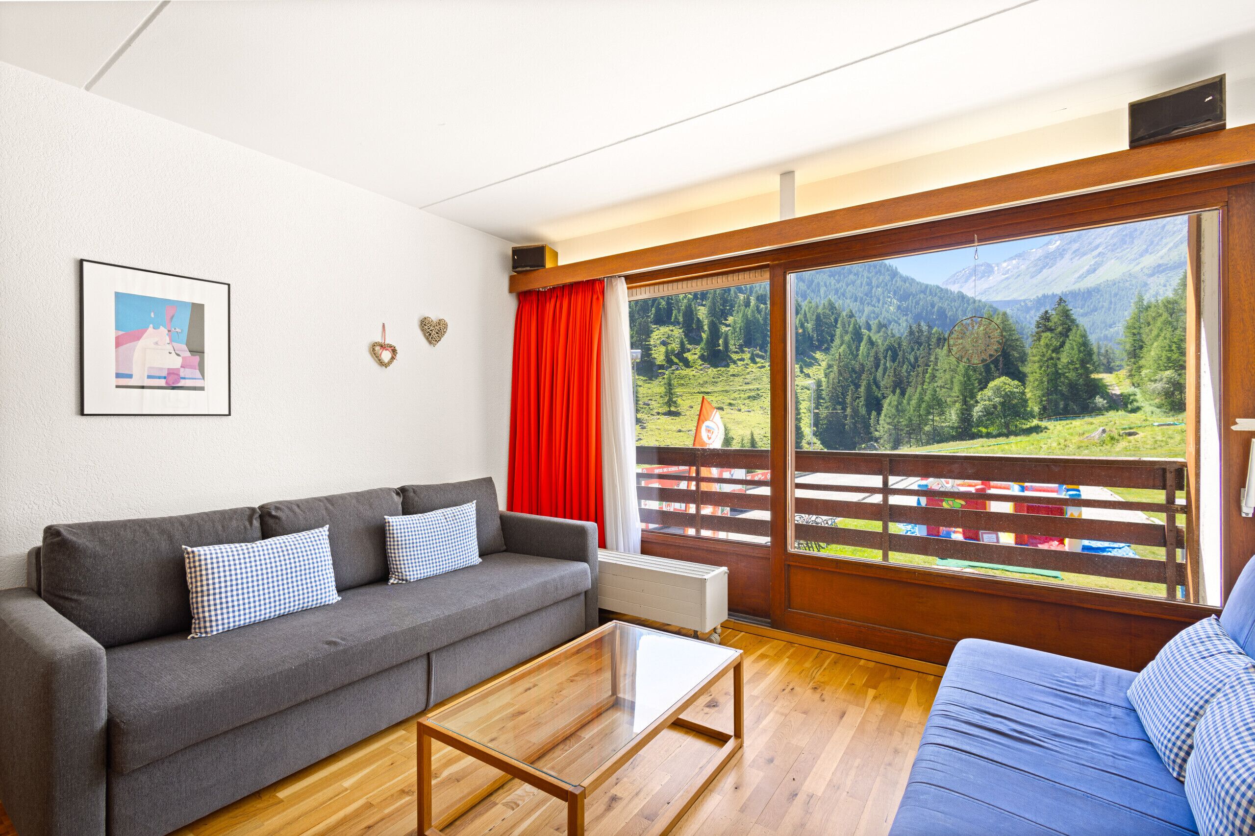 Ein Wohnzimmer mit grauem Sofa, blauem Sofa, Couchtisch aus Holz, großem Fenster mit Bergblick und einem gerahmten Bild an der Wand.