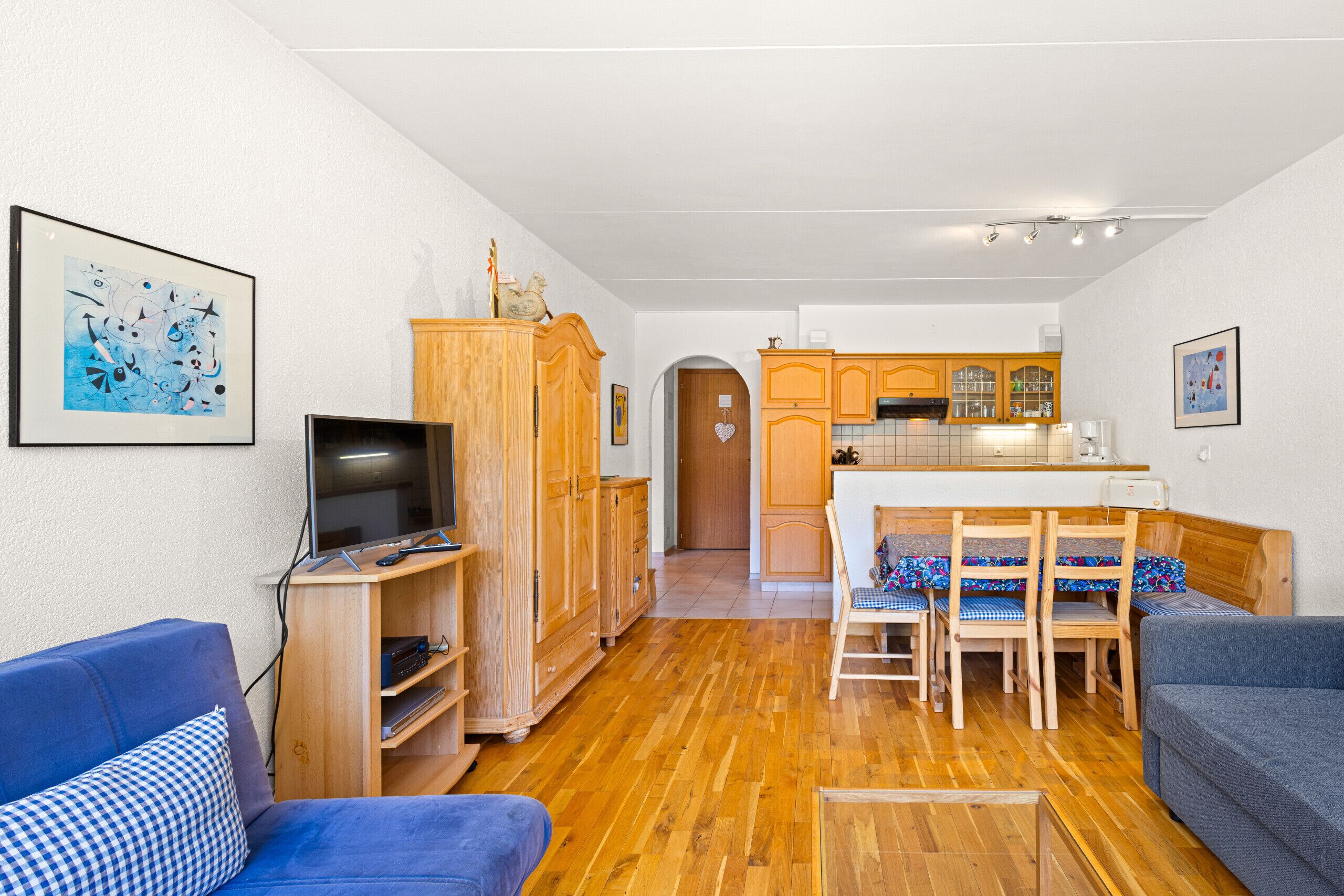 Un salon avec un téléviseur, des meubles en bois, un coin repas avec une table et des chaises, et une cuisine à l’arrière-plan.