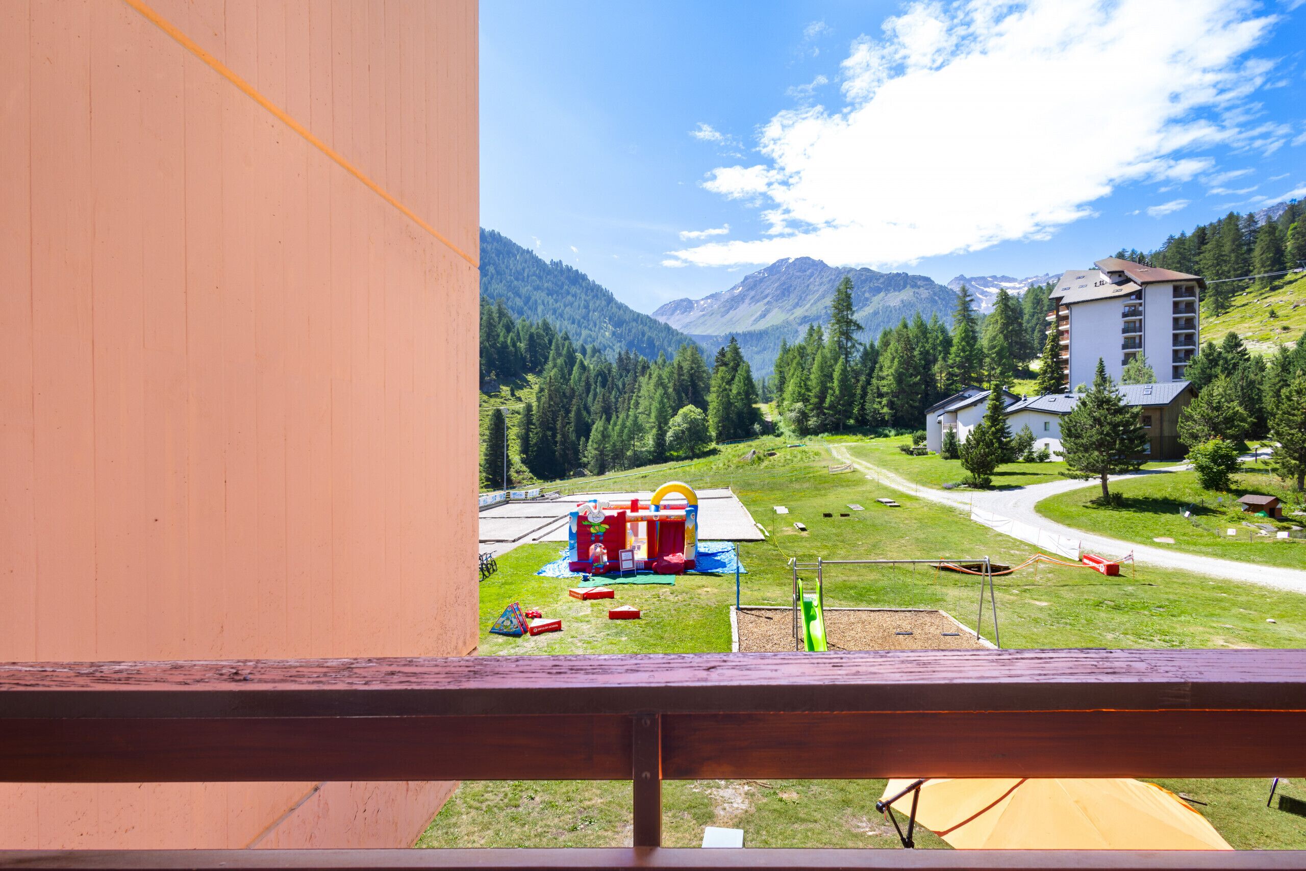 A view from a balcony overlooking a playground with play structures, a green field, and a mountain backdrop.