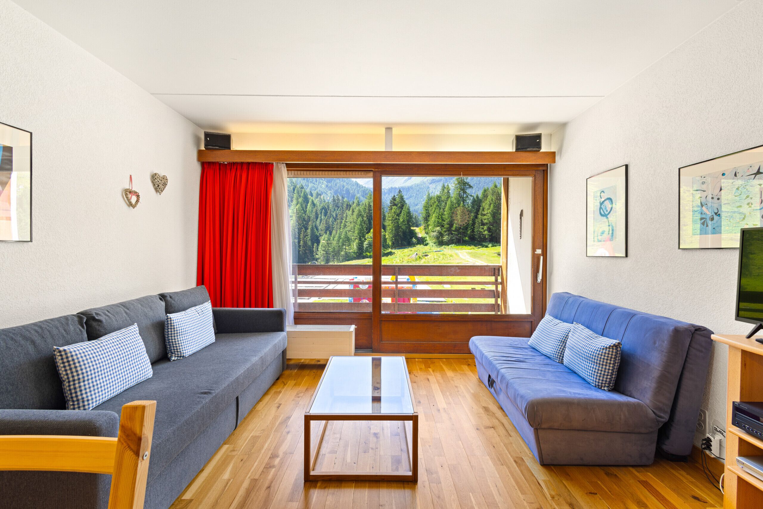 A living room with two sofas, a coffee table, and a large sliding glass door leading to a balcony with a view of a forested area.