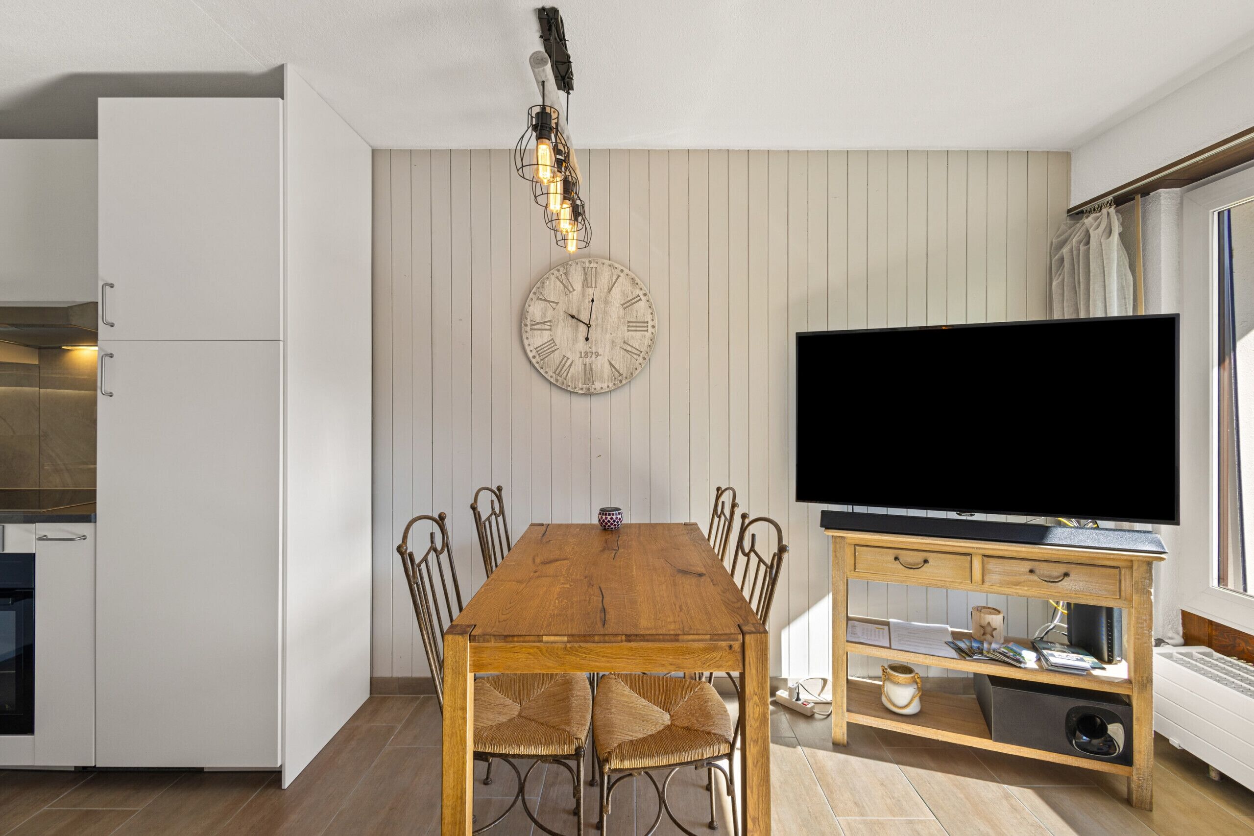 Un comedor con una mesa y sillas de madera, un reloj montado en la pared, un televisor sobre un mueble de madera y una zona de cocina con armarios blancos.
