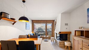 A cozy living room with a wooden dining table, chairs, a sofa, a fireplace, and a large window offering a mountain view. - Les Hauts-de-Nendaz B L7 - 4 pers - ski In/Out (Haute-Nendaz)