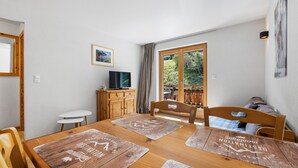 Uma área de refeições com mesa de madeira, cadeiras, uma televisão em um armário de madeira e vista para o verde pela janela.