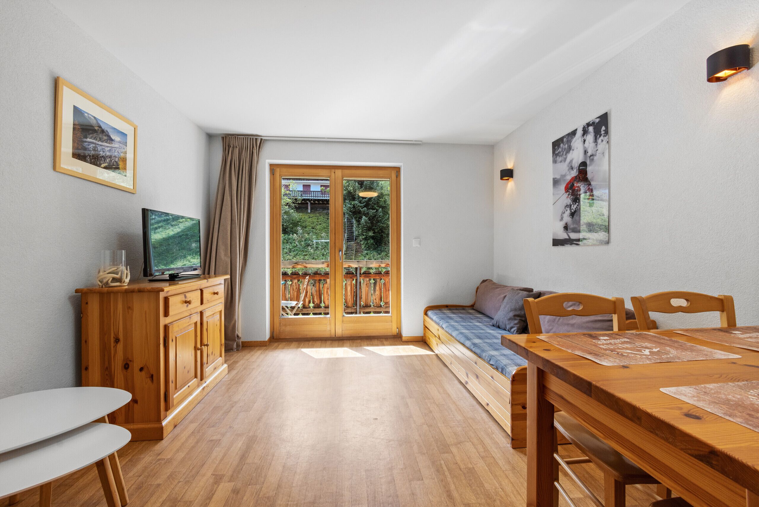A room with a wooden dining table, a sofa, a TV on a wooden cabinet, and a balcony with a view of greenery.