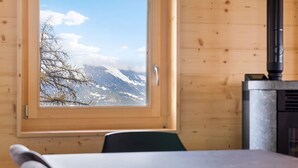A wooden cabin with a window offering a view of snow-capped mountains and a tree.