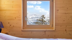 A wooden cabin room with a bed and a window overlooking a snowy mountain landscape.
