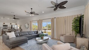 Ein geräumiges Wohnzimmer mit einem Ecksofa, einem runden Couchtisch und einem beigefarbenen Sessel. Es gibt Deckenventilatoren, ein großes Fenster mit Vorhängen und einen Blick ins Freie.