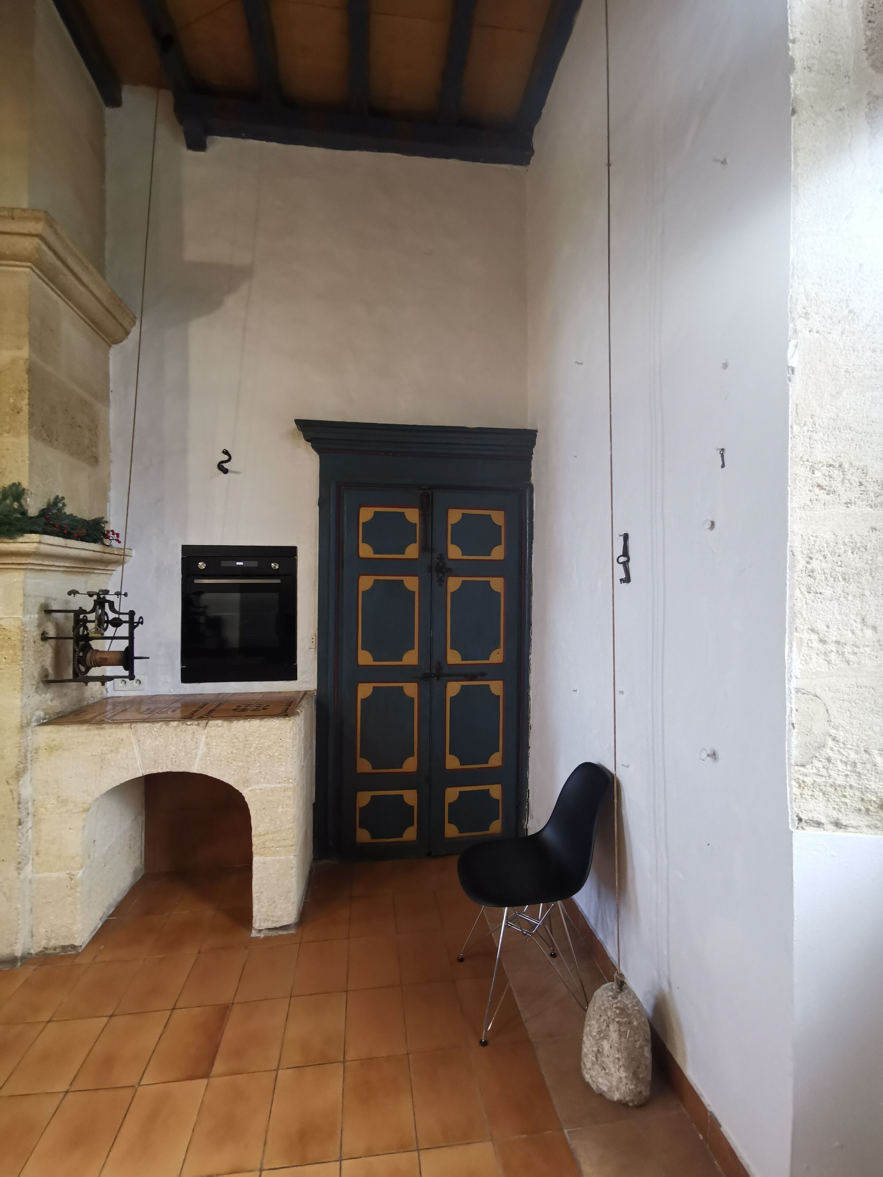 A narrow corridor with a tiled floor, a black door with decorative panels, a black chair, and a stone bench.