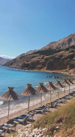 A beach with lounge chairs and thatched umbrellas, rocky hills in the background, and a clear blue sea. - Modern apartment Oleandra, 5 only 250 meters from the sea (Baska)