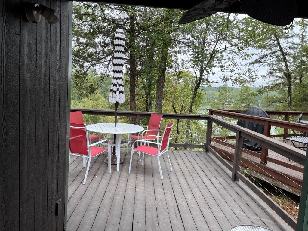 A wooden deck with a round table and red chairs, overlooking a forested area.