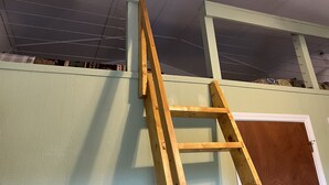 Una habitación con una escalera de madera apoyada en una pared, una puerta y un techo con cableado a la vista.