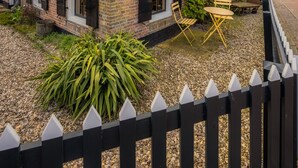Een bakstenen huis met een pannendak, een zwart hek van planken en een kleine tuin met een houten stoel en tafel.