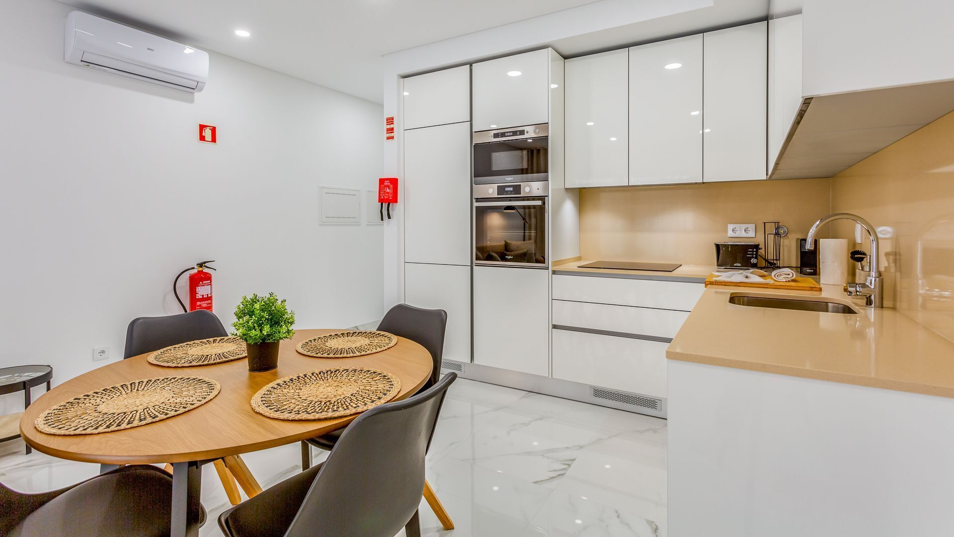 A modern kitchen with white cabinets, a built-in oven, and a sink. There is a dining area with a wooden table and chairs, and a small plant on the table.