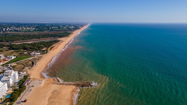An aerial view of a coastal area with a sandy beach, clear blue waters, and a residential area with buildings and greenery. - Sublime and Bright 2 BDR Apartment by LovelyStay (Quarteira)