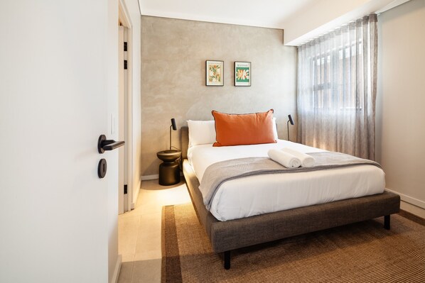 A neatly made bed with a grey headboard, a brown throw pillow, and a white blanket. There are two framed pictures on the wall, a small round table with a lamp, and a window with sheer curtains. - Tyne Aparthotel l Steam Room l Pool (Umhlanga)