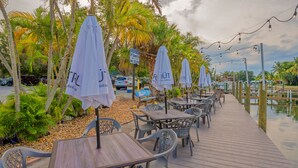 An outdoor dining area with tables and chairs, umbrellas, and a wooden deck overlooking water. - ALL-NEW Waterfront with Balcony, Pool, Tiki Bar, Gym (Sarasota)