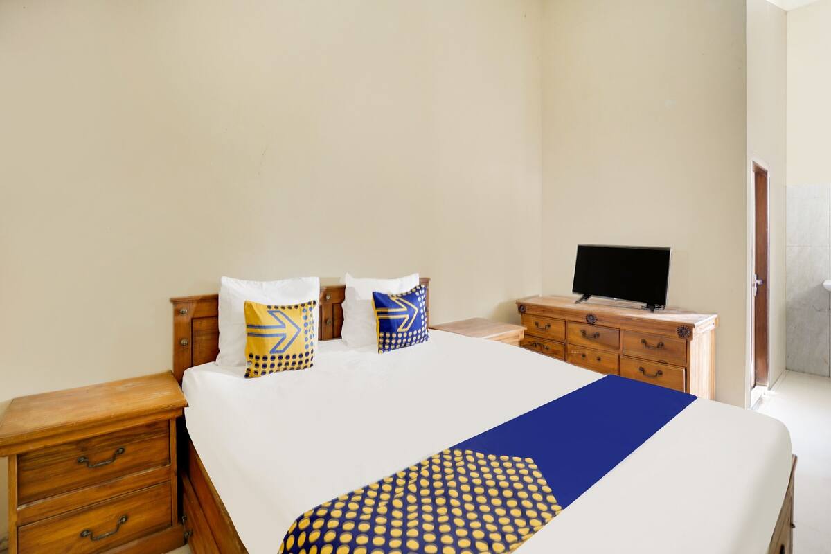 A neatly made bed with a blue and yellow patterned bedspread, two pillows with geometric designs, a wooden nightstand with drawers, a wooden dresser with a flat-screen TV on top, and a door leading to another room.