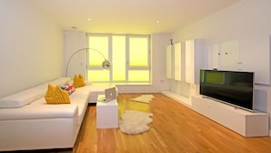 A modern living room with a white sofa, a flat-screen TV, and a large window with natural light. - Penthouse Kew (Brentford)