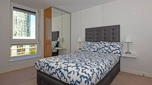 A bedroom with a large bed, two bedside tables with lamps, a window with a view of buildings, and a ceiling light. - Penthouse Kew (Brentford)