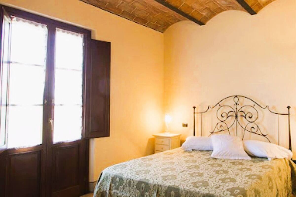 Un dormitorio con cama, mesita de noche, ventana y lámpara fijada en la pared.