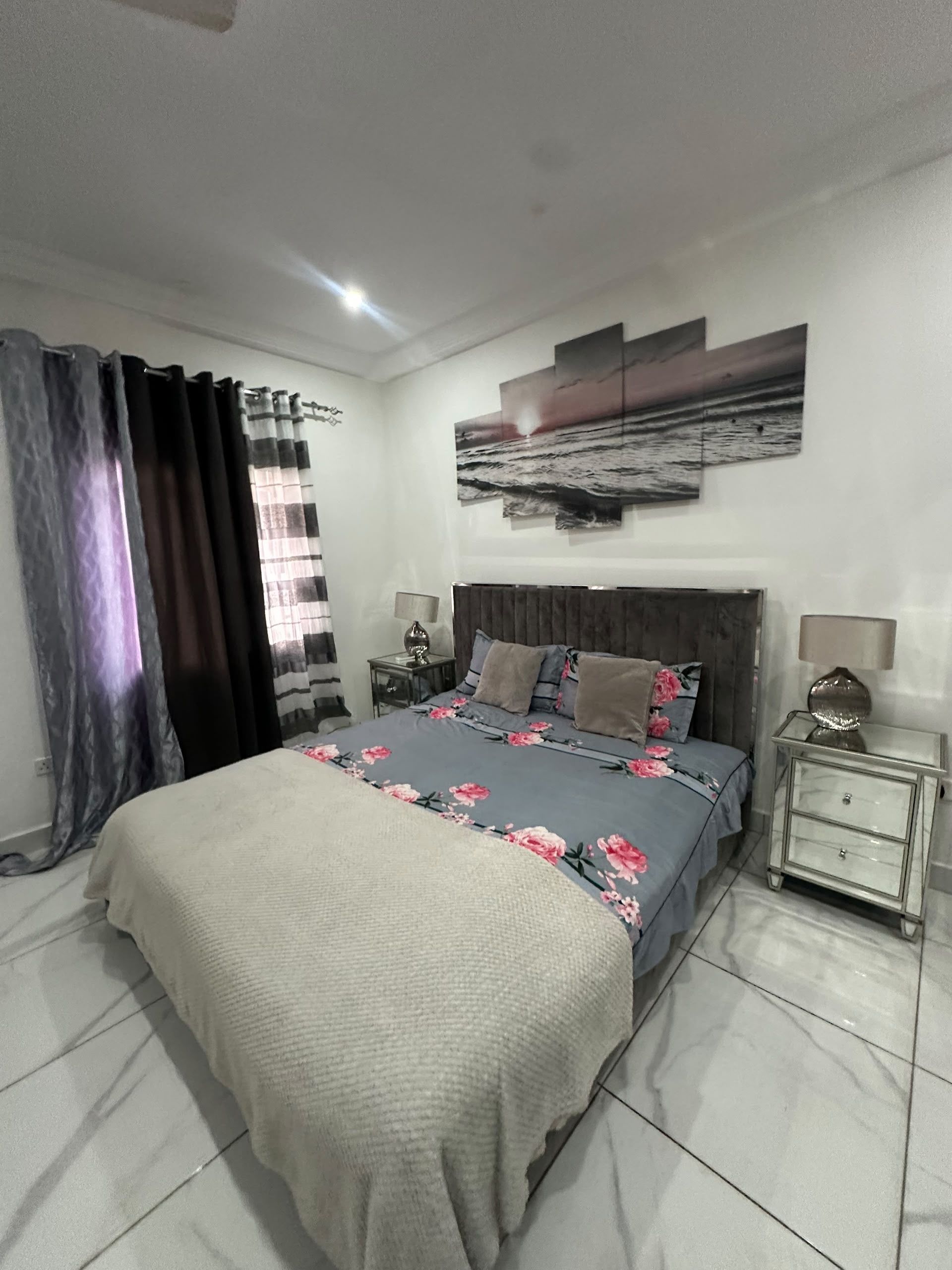 Un dormitorio con cama, cortinas, mesita de noche, lámpara y una obra de arte colgada en la pared.