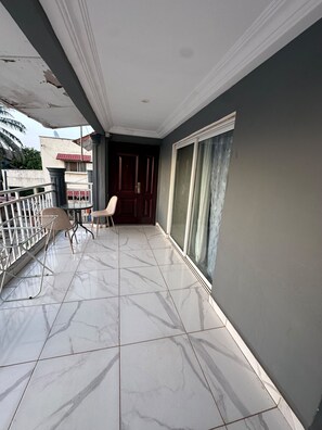 Un balcón con suelo de baldosas, una silla, una mesita y una puerta que da a una habitación interior.