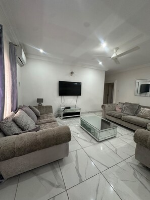 A modern living room with two sofas, a glass coffee table, a flat-screen TV mounted on the wall, and a ceiling fan. - Luxury Spacious two bedroom apartment for comfortable living. Well furnished . (Banjul)