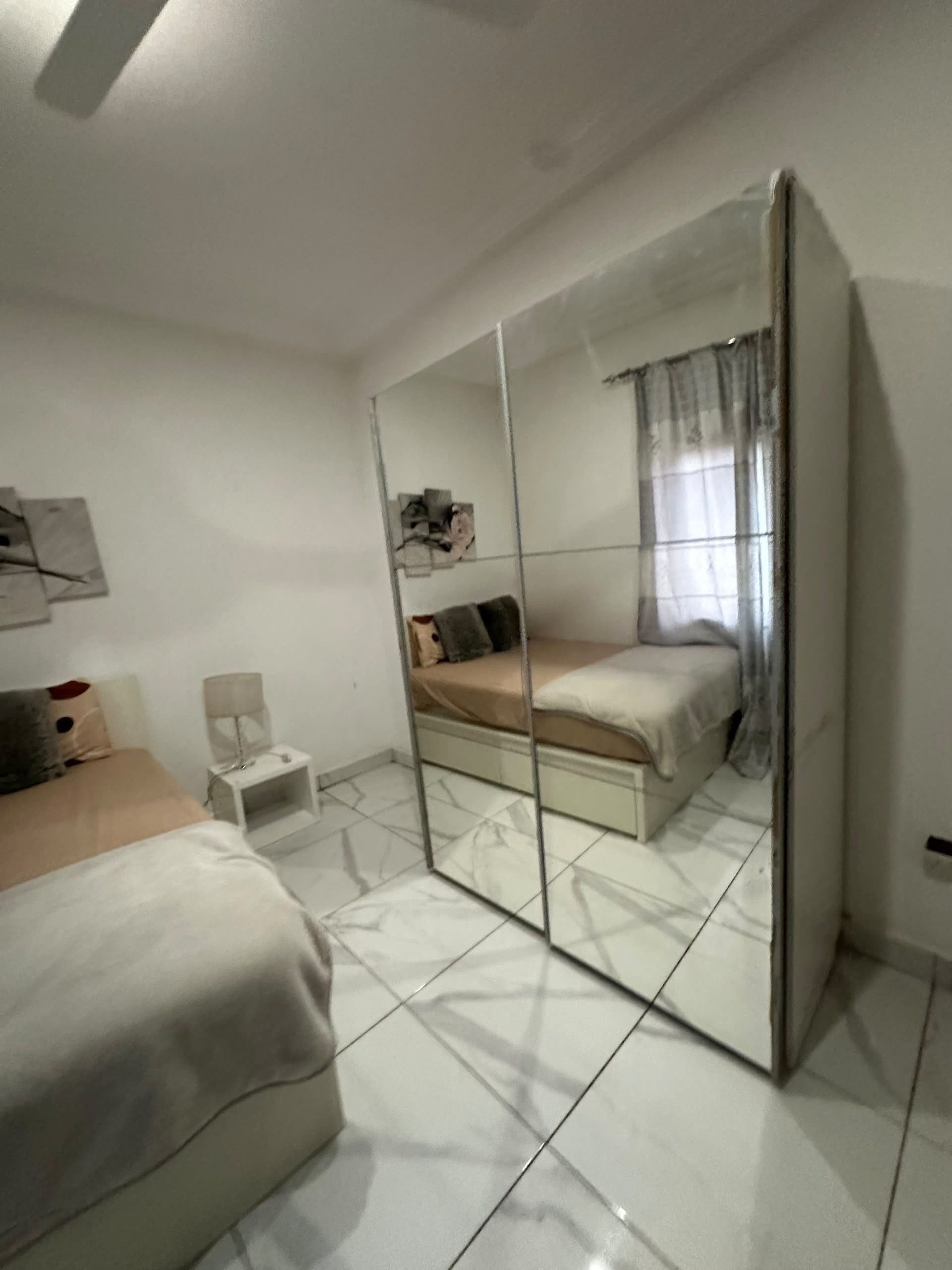 A bedroom with a bed, a small bedside table, a chair, a mirror, and a window with curtains.