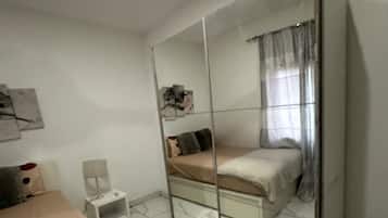 A bedroom with a bed, a small bedside table, a chair, a mirror, and a window with curtains.