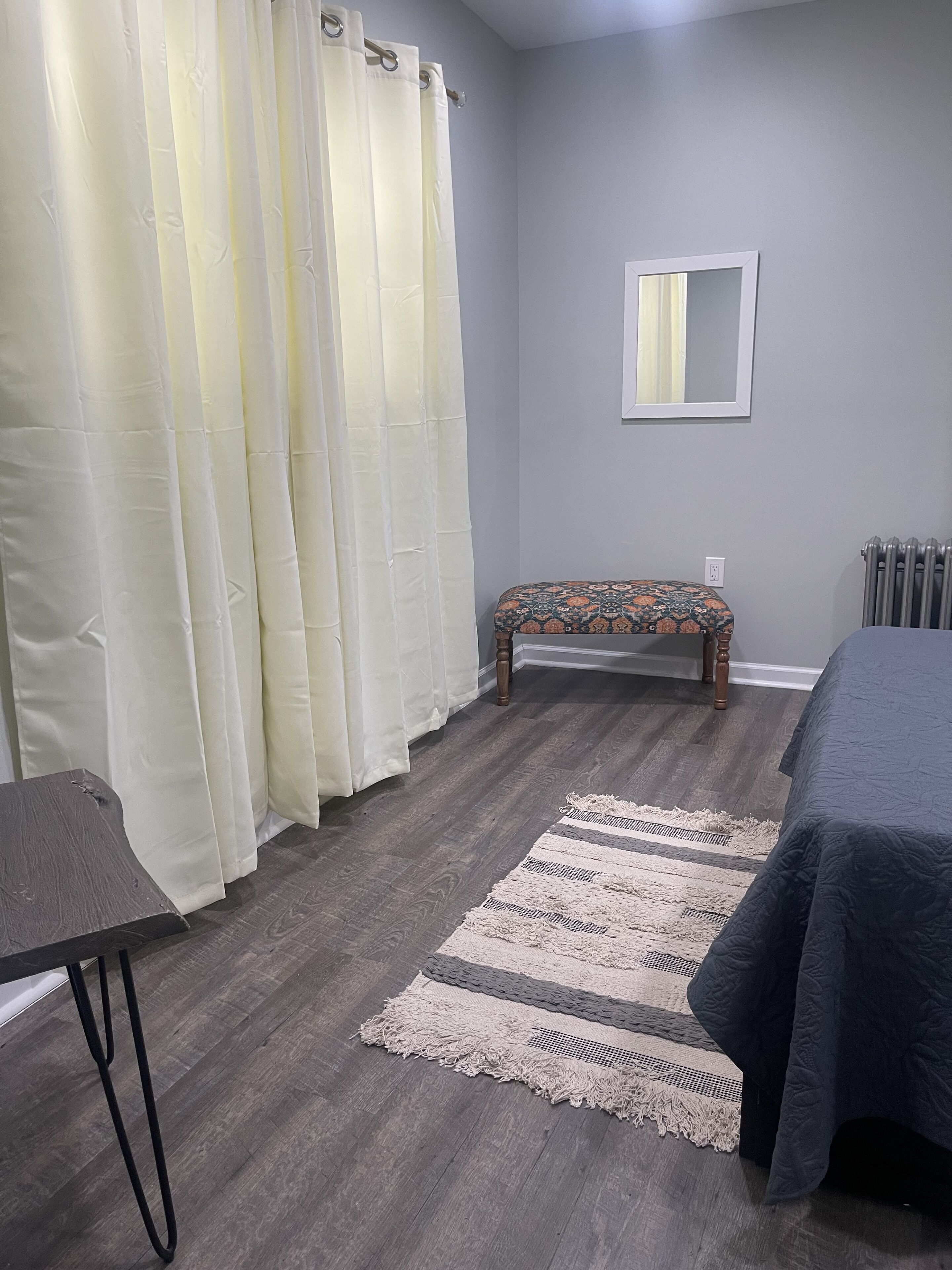 A bedroom with a bed, a bench, a mirror, a radiator, and a curtain.