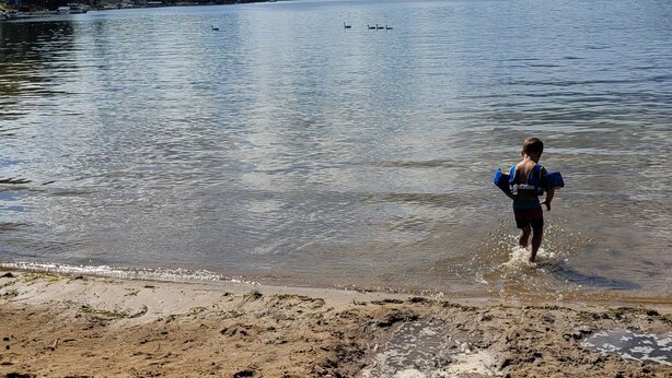 Une plage où une personne court vers l’eau, un rivage sablonneux et un lac où l’on aperçoit des bateaux au loin.