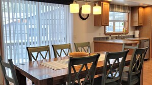 Un coin repas avec une table et des chaises en bois, une cuisine à l’arrière-plan et une fenêtre avec des stores.