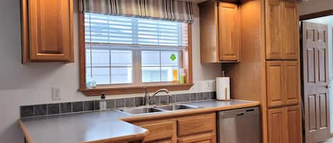A kitchen with wooden cabinets, a stainless steel dishwasher, and a window with blinds.
