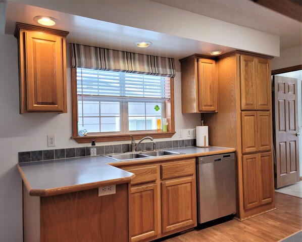 A kitchen with wooden cabinets, a stainless steel dishwasher, and a window with blinds. - 13-person home for families & groups! (Lake)