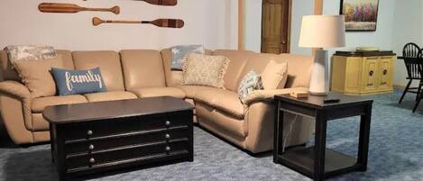 A living room with a beige sectional sofa, a dark wooden coffee table, a small side table with a lamp, and a wall-mounted paddleboard display.