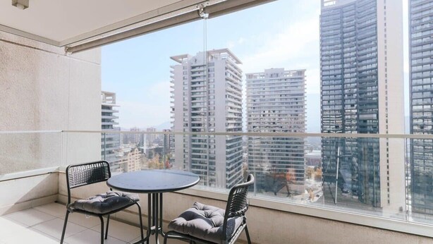 A balcony with two chairs and a table, overlooking a cityscape with high-rise buildings. - Boho Apartment - Pool, Parking & Mall. (Las Condes)