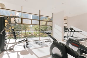 A modern gym with cardio equipment, weight machines, and a large window offering a view of greenery and buildings. - Boho Apartment - Pool, Parking & Mall. (Las Condes)