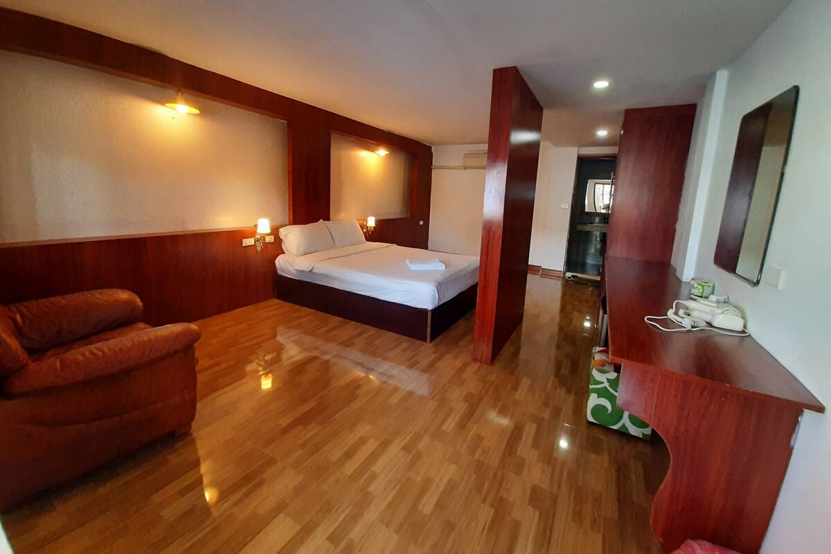 A hotel room with a bed, a wooden chair, a desk with a mirror, and a small kitchenette area.
